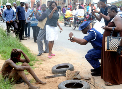 By standers, including a Civil Defence officer take snaps of the alleged ritualist at Ring Road, Ibadan 