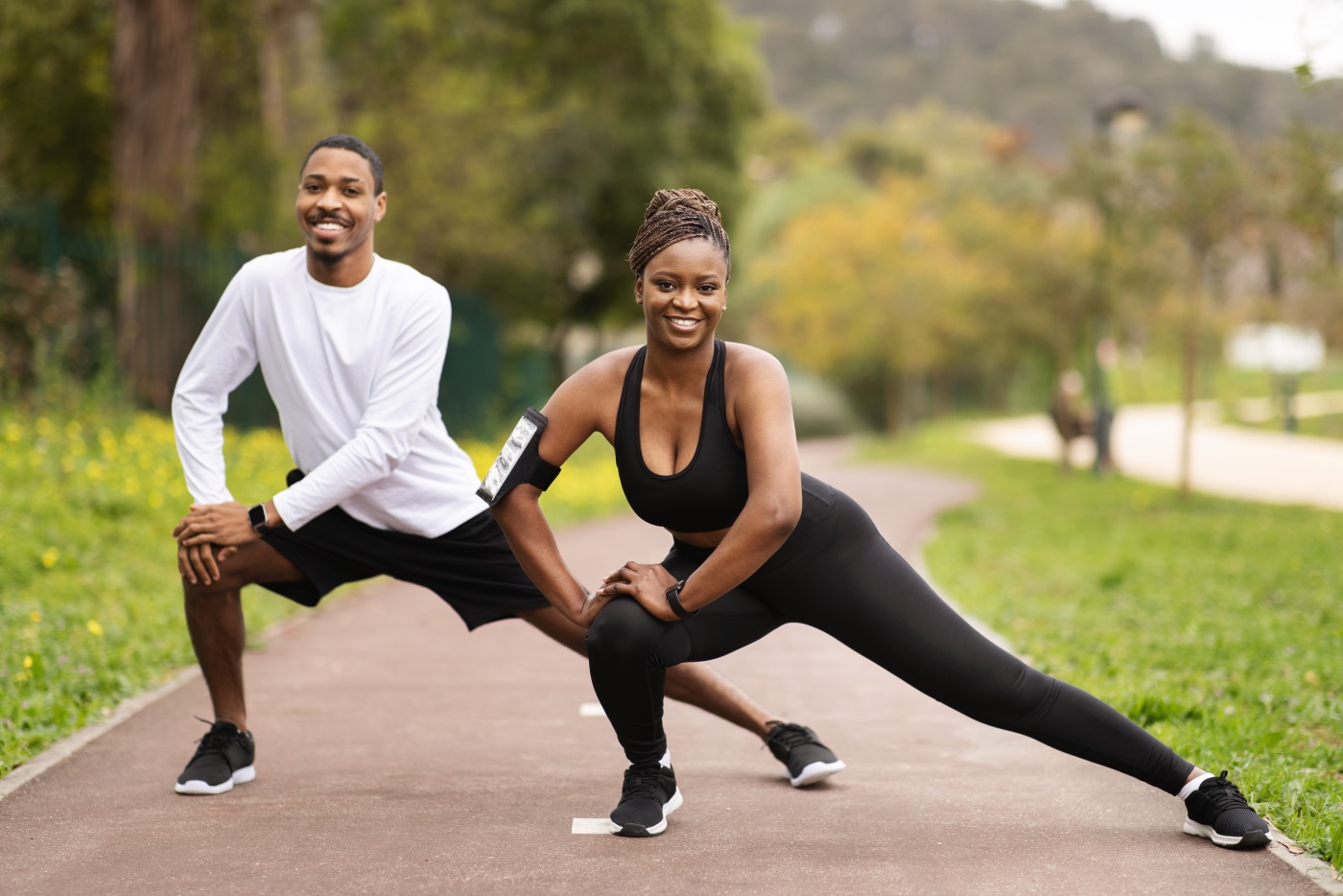 Cheerful young black couple with phone doing leg exercises in summer, enjoy fit. Warm-up before running, sports, outdoor training, exercises for body care and relationships