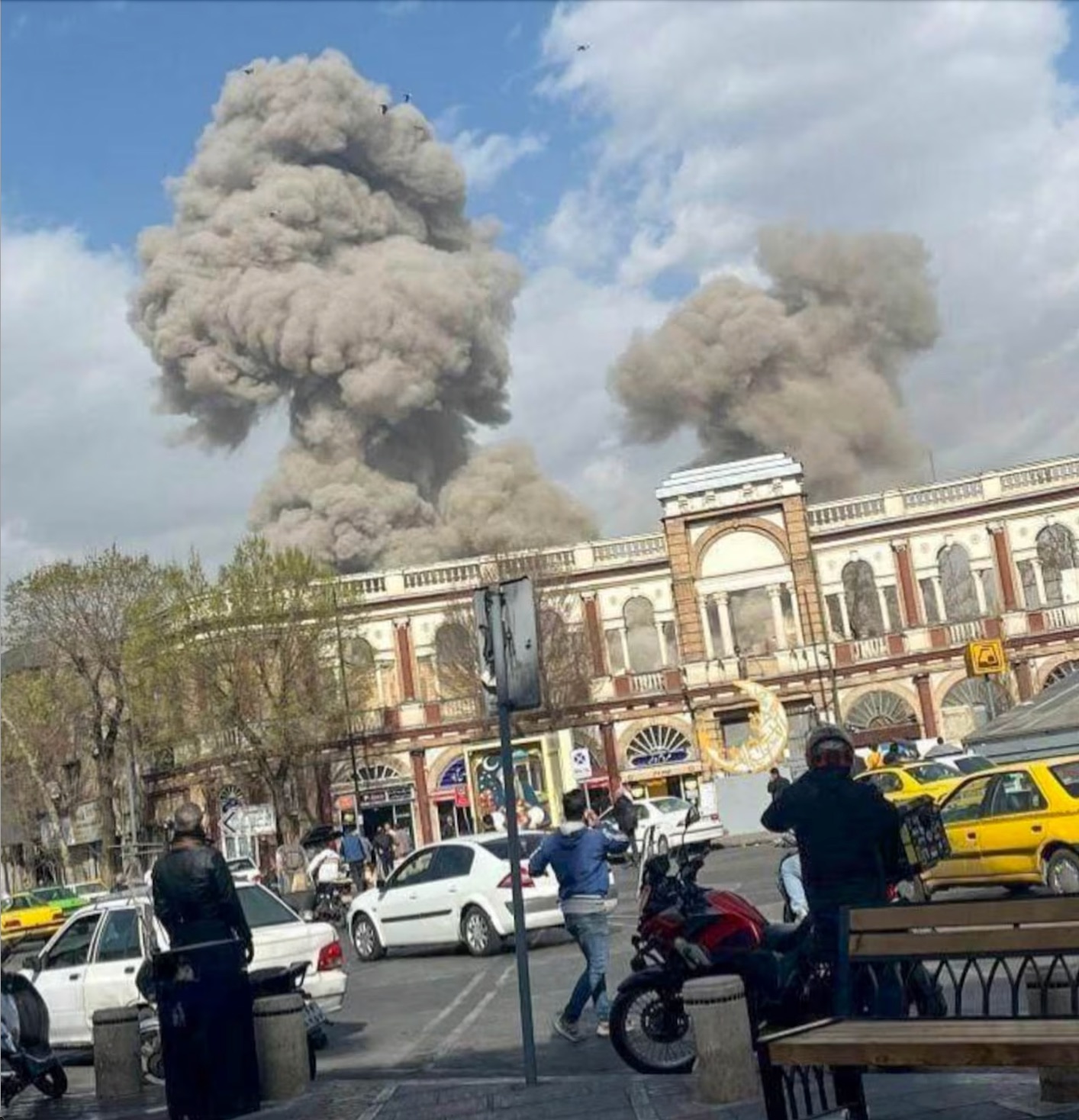 Onlookers stand on the street as smoke rises over buildings in the city. Noor Pictures/Shutterstock